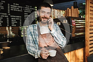 Portrait of young barista guy talking on cellphone in street cafe or coffeehouse outdoor