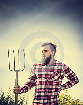 Portrait of young bearded farmer in red checkered shirt with old pitchfork on sky nature backgrund, toned photo