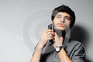 Portrait of young bearded guy shaving beard with trimmer. On textured background of grey.