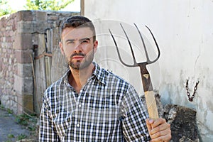 Portrait of young bearded handsome farmer in casual checkered shirt with old pitchfork on rustic background