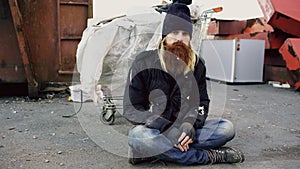 Portrait of young bearded homeless man sitting on a sidewalk near shopping cart ang garbage container during cold winter