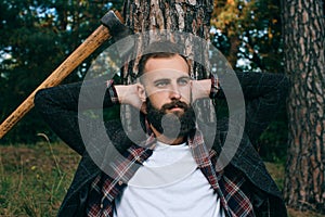 Portrait of a young bearded lumberjack resting near a tree in the woods