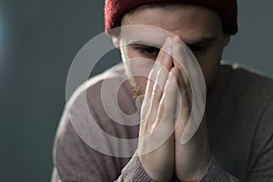 Portrait of a young bearded man, hands clasped on his face, prays, dropping his eyes down. Studio photo on a gray background