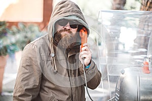 Portrait of young bearded man using public phone wearing hoodie jacket and black sunglasse