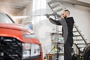 Portrait of of young bearded man worker washing red car under high pressure water