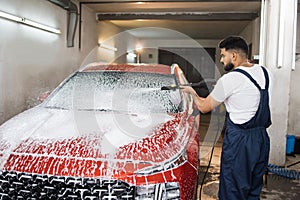 Portrait of young bearded man in working clothes, car wash employee, washing the car windshield