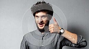 Portrait of young bearded satisfied guy with disheveled hair, shows thumbs up on background of grey.