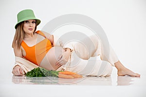 Portrait of young beauteous pregnant woman with long dark hair wearing green bucket hat, lying near bunch of carrots.