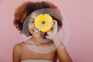 Portrait of a young beautiful African-American woman closes one eye with a yellow flower, looks at the camera and smiles