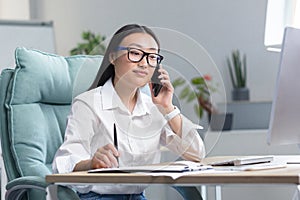 Portrait of a young beautiful Asian business woman in glasses in a white shirt talking on the phone