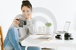 Portrait of young beautiful Asian female blogger photographer sitting at modern office while checking previews on camera.Happy