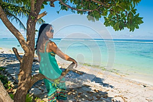 Portrait of young beautiful asian girl sitting in shade under the tree on tropical beach looking at ocean