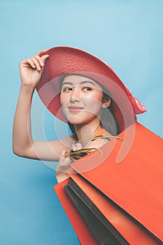 Portrait of young beautiful asian girl wearing dress and sun hat holding shopping bags and smile isolated over blue background.
