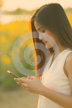 Young beautiful Asian woman using mobile phone against field of blooming sunflowers