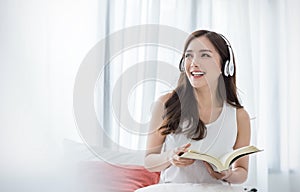 Portrait of young beautiful asian woman relax holding book listen to music from headphone in bedroom.