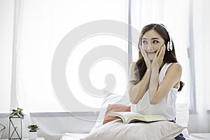 Portrait of young beautiful asian woman relax holding book listen to music from headphone in bedroom.