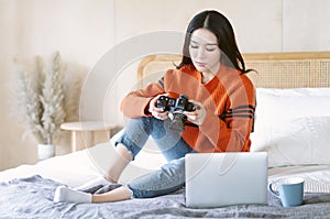 Portrait of young beautiful Asian woman in warm knitted clothes with professional camera and checking picture while sitting on bed
