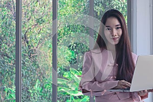 Portrait of young beautiful and attractive Asian business woman with smiling in pink dress hold labtop in modern officeas a