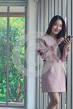 Portrait of young beautiful and attractive Asian business woman with smiling in pink dress hold smart phone in modern officeas a