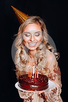 Portrait of a young beautiful blond girl holding birthday cake with candles on black background