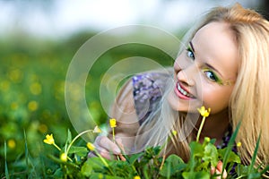 Portrait of young beautiful blond woman lying in grass and flowers on summer day with green nature landscape