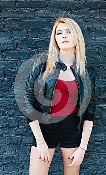 Portrait of a young beautiful blonde girl in a black jacket and shorts posing near brick wall