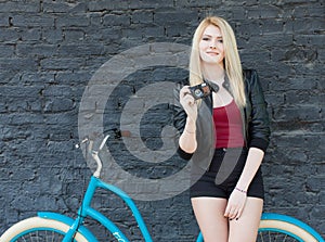 Portrait of a young beautiful blonde girl in a black jacket and shorts posing near the brick wall next to a bright blue vintage bi