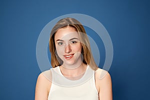 Portrait of young, beautiful, blonde girl posing, looking at camera with smile against blue studio background. Happiness