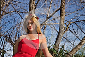 Portrait of young, beautiful, blonde woman with a red dress and flower tiara, leaning on a wooden railing, calm and relaxed.