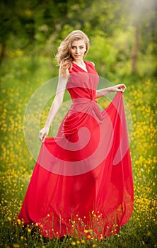 Portrait of young beautiful blonde woman wearing a long red elegant dress posing in a green meadow. Fashionable attractive