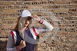 Portrait of young, beautiful, blonde woman, with white cap, and baseball jacket, in sensual and flirtatious attitude, with a wall