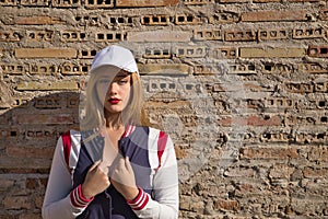 Portrait of young, beautiful, blonde woman, with white cap, and baseball jacket, in sensual and flirtatious attitude, with a wall