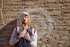 Portrait of young, beautiful, blonde woman, with white cap, and baseball jacket, in sensual and flirtatious attitude, with a wall