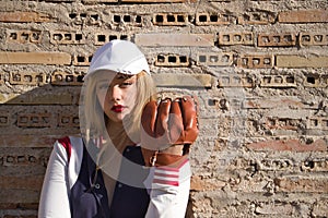 Portrait of young, beautiful, blonde woman with white cap, jacket, baseball glove and ball, leaning against a broken brick wall in