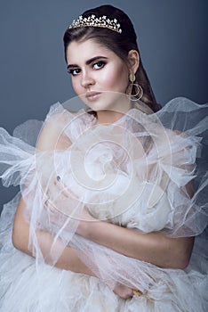 Portrait of young beautiful bride in diadem and earrings with naked shoulders pressing skirt of her wedding dress to her breast