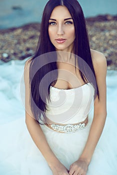 Portrait of young beautiful bride with long black hair in wedding dress sitting at the seaside and looking straight tenderly photo