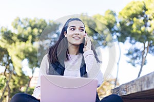 Portrait of a young beautiful brunette sitting on park with laptop while using smartphone to call