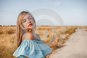 Portrait of a young beautiful caucasian blonde girl in a light blue dress standing on a field with sun-dried grass next to a small