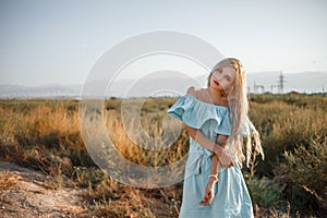 Portrait of a young beautiful caucasian blonde girl in a light blue dress standing on a field with sun-dried grass next to a small