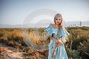 Portrait of a young beautiful caucasian blonde girl in a light blue dress standing on a field with sun-dried grass next to a small