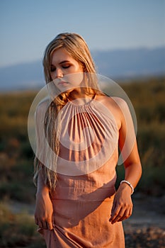 Portrait of a young beautiful Caucasian blonde woman in a pink dress in a deserted field against the sunset