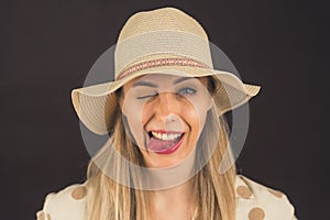 Portrait of a young beautiful caucasian blonde woman in studio, sticking out tongue, wearing beige hat over black or