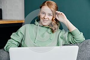 Portrait of a young beautiful caucasian woman dressed in a green hoodie working at home using a laptop