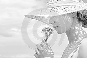 Portrait of young beautiful cheerful woman wearing straw sun hat, smelling small bouquet of yellow wild florets, against