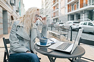 Portrait of young beautiful fashionable blond woman in warm clothes sitting in an outdoor cafe with laptop computer, drinking cup