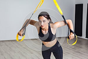 Portrait of young beautiful fit woman in black sportswear training arms with trx fitness straps in the gym doing push ups train