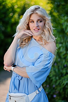 Portrait of a young beautiful girl 21 years old in a blue summer dress with a white belt bag. The woman smiles