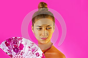 Portrait of a young beautiful girl with bright make-up and colored fan in hands close-up on the pink background. Geisha