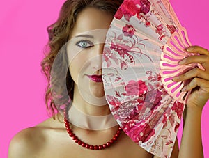 Portrait of a young beautiful girl with bright make-up and a fan in hands close-up on the pink background