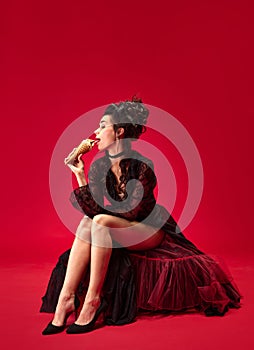 Portrait of young beautiful girl in elegant black dress sitting, eating ice cream against red background